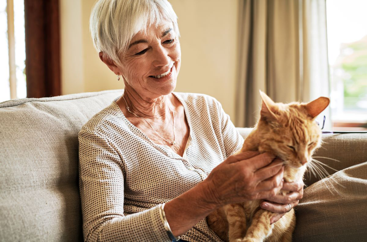 Mujer mayor sonriendo y acariciando a un gato en un sofá.